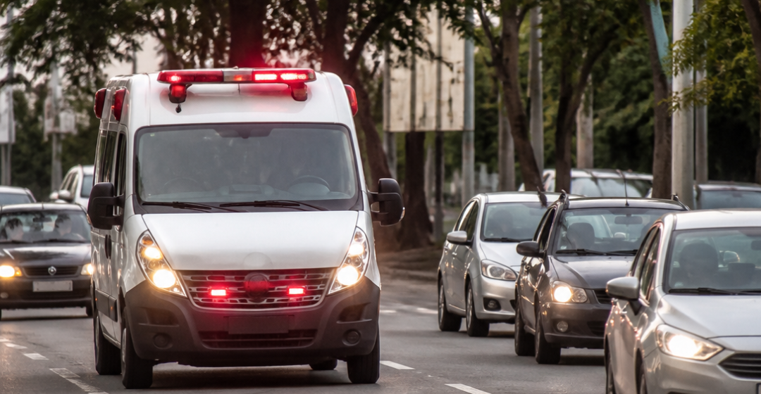 Ambulância com sirene e luzes de emergência trafegando enquanto veículos abrem passagem em uma avenida urbana brasileira