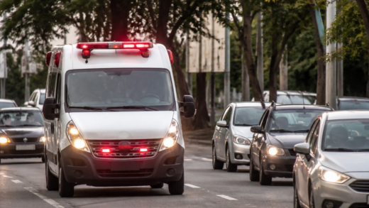 Ambulância com sirene e luzes de emergência trafegando enquanto veículos abrem passagem em uma avenida urbana brasileira